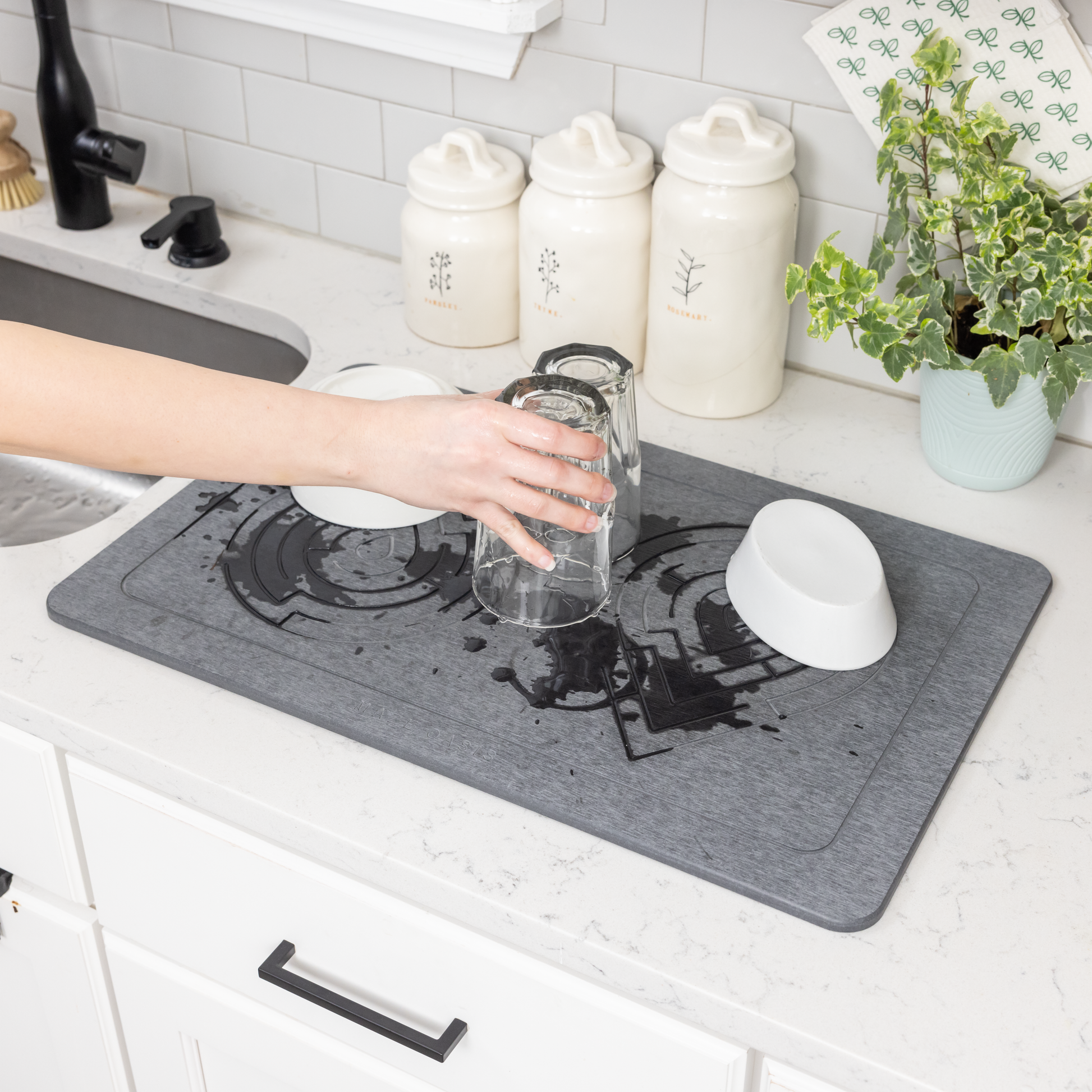 A diatomite stone mat in a gray color placed on a kitchen counter, with a person pouring water from a glass onto it, demonstrating its quick-drying feature.